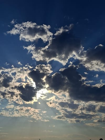 The image depicts a dynamic sky over Казашко, Bulgaria, characterized by a dramatic interplay of sunlight and clouds. The sky is a deep blue, transitioning to lighter hues near the horizon. Scattered across the sky are numerous cumulus clouds, some brightly illuminated by the sun, appearing white and puffy, while others are darker, casting shadows and suggesting depth. Rays of sunlight pierce through gaps in the clouds, creating visible beams of light that add to the atmospheric effect. The lower portion of the image shows a faint, hazy horizon, with minimal details of the landscape or any structures. There are no people or specific identifiable objects in the foreground or midground. A single bird is visible in flight in the lower right quadrant of the image, silhouetted against the lighter sky. The overall impression is one of a partly cloudy sky with the sun partially obscured.