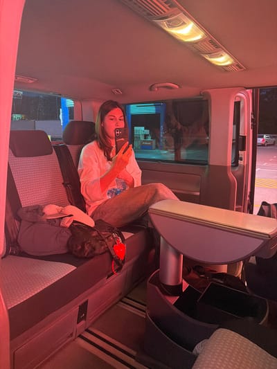 A woman is in the backseat of a vehicle, possibly a van, in an urban setting. She is holding a smartphone up to her face, presumably taking a photograph or video. She is wearing jeans and a light-colored shirt. Her hair is brown. The vehicle's interior is lit with warm-toned lights. A tray table is folded out in front of her. A bag is on the seat next to her. Through the windows, signage for "O-Drive" and "Petrol" are visible, suggesting the location is a petrol station. The red lighting casts an odd tone and shadows throughout. Without further evidence it is impossible to locate the city of Stara Pazova Municipality, Serbia.