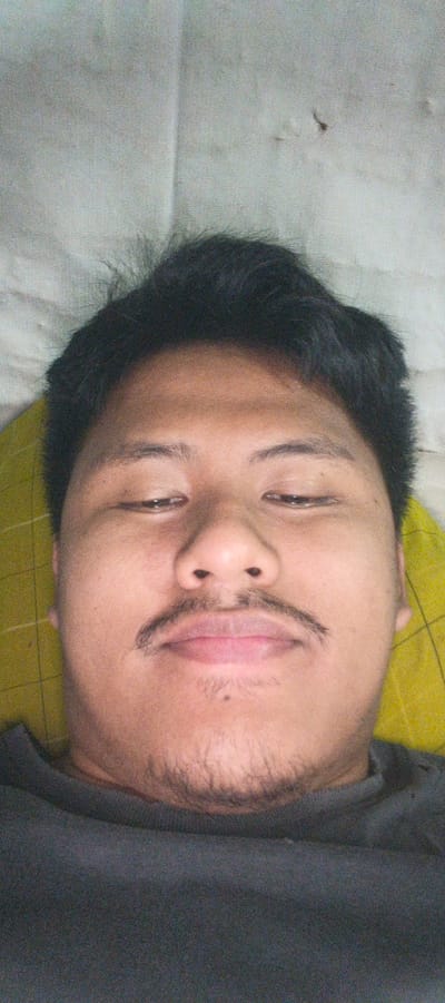 A close-up, overhead selfie shows a person lying down, possibly in Kemuning, Indonesia. The person has dark hair and is wearing a dark gray shirt. Their eyes are partially closed, and they have a mustache and a light beard. The background consists of a white, textured surface and a yellow patterned object, likely a pillow or blanket.