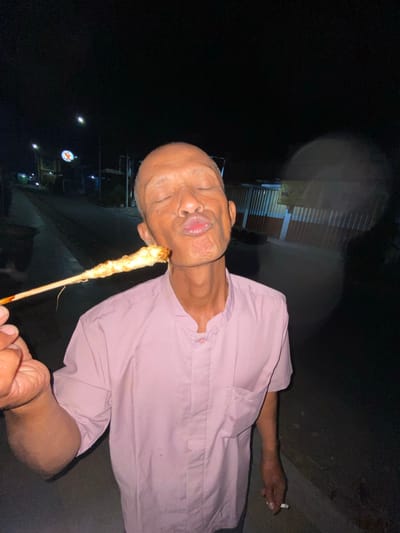In Tanggul, Indonesia, an elderly bald man with closed eyes and pursed lips is shown outdoors at night, holding a skewer of grilled food to his mouth as if to kiss it. He is wearing a light pink button-up shirt. The background shows a dimly lit street with streetlights, buildings, and a fence. The man is also holding a cigarette in his left hand.