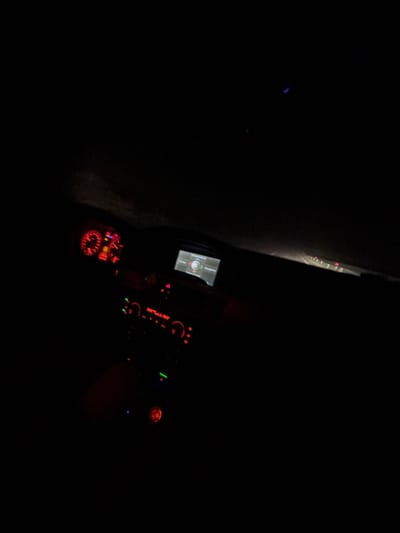 The image depicts the interior of a car at night. The dashboard is illuminated with red and green lights. A navigation screen is visible. The car appears to be in motion on a road, based on the blurry light streaks outside the window. The location is possibly Rahovtsi, Bulgaria.