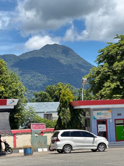 The image depicts a scene in Sukamulia, Indonesia, with a mountainous backdrop. In the foreground, a white van is parked in front of a building with a blue facade and red trim. The building has signage for "BSI BANK SYARIAH INDONESIA ATM." To the left of the van, there is a gas station canopy with red and white detailing and a red sign that reads "AIR & ANGIN." A person on a motorcycle is pulling away from the gas pump area. Behind the gas station, there's a wall and a cluster of trees. The mountain in the background is covered in lush green vegetation, and the sky is a clear blue with scattered white clouds. The overall impression is a typical day at a service station in a tropical or subtropical region, with a prominent natural landscape feature.