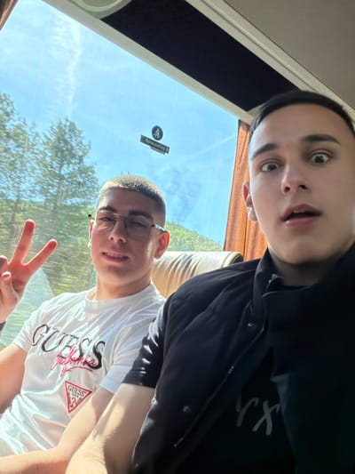 Two young men are in a vehicle, possibly a bus, as suggested by the window view showing trees and a "Notausstieg" emergency exit sign in German. The man closest to the camera is wearing glasses and a white Guess t-shirt, flashing a peace sign. The other man is wearing a black jacket. The location in Gorno Aleksandrovo, Bulgaria, cannot be independently confirmed.