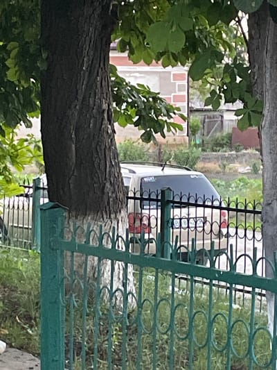 The image depicts a residential area in Tovstoluh, Ukraine. A large tree trunk, painted white at its base, stands prominently in the foreground, partially obscuring a green metal fence. Behind the fence, a light-colored SUV is visible, with its rear end facing the camera. The vehicle has a license plate with Cyrillic characters and numbers that read "BB 42 CM". The background shows a building with decorative brickwork and a rustic fence enclosing a yard with overgrown vegetation. The overall scene suggests a quiet, possibly rural or suburban setting.