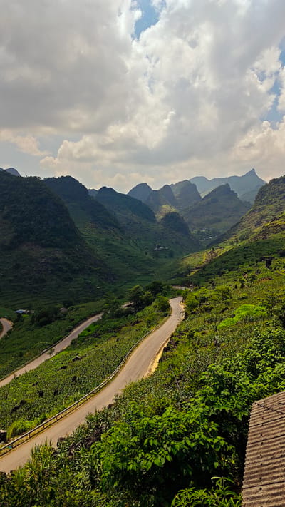 The image shows a winding road cutting through a mountainous region in Lũng Phìn Commune, Vietnam. The road is asphalt and has metal guardrails. The mountains are covered in lush, green vegetation. Some buildings are visible in the distance. The sky is cloudy. The view is from a high vantage point. A section of a corrugated roof is visible in the lower right corner.