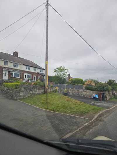The image depicts a residential street scene in Halkyn, United Kingdom, taken from inside a vehicle. The perspective is slightly tilted. On the left side of the street, there are terraced houses constructed from brick and rendered walls. The roofs are pitched and predominantly dark brown. A wooden utility pole with multiple power lines is visible, crossing the street diagonally. On the right side of the street is a stone wall bordering properties, with various bushes and plants growing above it. Several wheelie bins are situated near the wall. The road surface is gray asphalt with some visible water marks. The sky is overcast and gray. The interior of the vehicle is visible on the right side of the image, with a portion of the window frame in view.