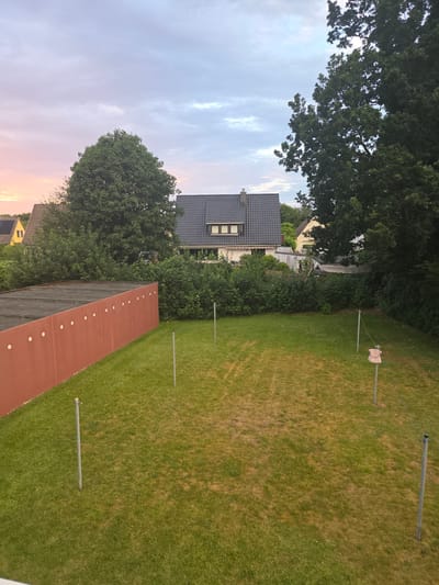 The image depicts a residential backyard in Uetersen, Germany, at dusk. A red, concrete wall with circular indentations runs along the left side of the frame. The backyard itself is covered in grass, with several metal poles spaced across it, some of which appear to be clothesline supports. One pole in the middle right has a decorative, pink object resembling a dress or garment hanging from it. In the background, there are several houses with pitched roofs, surrounded by lush green trees and bushes. The sky is cloudy, with soft hues of pink and orange on the left side, suggesting the setting sun. The overall atmosphere is peaceful and suburban.