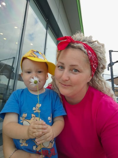 This is a selfie of a woman and a young child taken outdoors. The woman has curly blonde hair and is wearing a bright pink shirt and a red bandana. The child is wearing a yellow baseball cap and a blue t-shirt with cartoon characters on it. The child is holding a daisy up to their nose. They are positioned in front of a building with large windows, reflecting the sky and some lights. The overall setting suggests a casual outdoor environment, potentially in a public space like a cafe or park. The image does not provide specific visual cues to identify the location as Novoe Mart'yanovo, Russia.