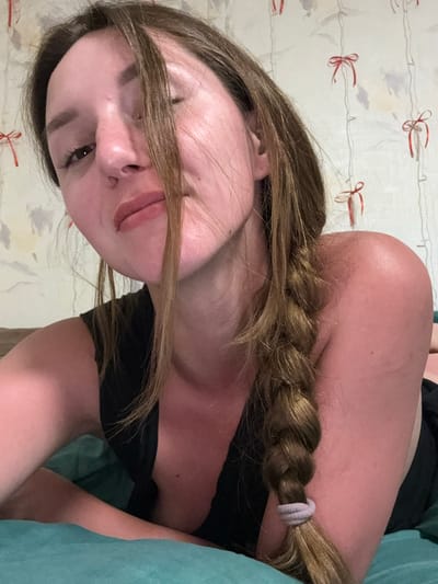 The image shows a close-up, low-angle shot of a young woman lying on her side on a teal-colored surface, possibly a bed or sofa. She has long, brown hair, mostly falling over her face, with a braid visible down her back. She is wearing a black top. Her skin appears reddish, suggesting a sunburn. The background is a wall with a patterned wallpaper featuring delicate red bows and vertical lines. The scene is indoors, and the lighting is soft. No specific location in Zhodzina, Belarus is identifiable from the image.