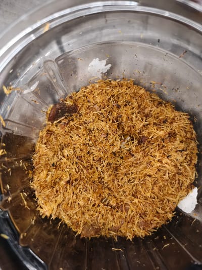 The image shows a close-up, overhead view of a pile of shredded, golden-brown material, likely food, contained within a clear plastic bowl. The material appears to be finely shredded and textured, with some darker brown pieces interspersed. The bowl is clear and ribbed at the bottom, and its rim is visible at the top and sides of the frame. There are a few small white flecks, possibly bits of salt or other ingredients, visible on the shredded material and on the inside of the bowl. The lighting is somewhat harsh, creating reflections on the plastic surface. There is no indication of the specific location within Barengkrajan, Indonesia, or any people, actions, or interactions depicted in the image.