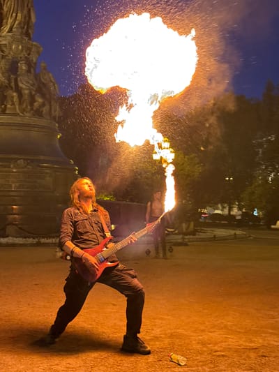 A man with long, reddish-brown hair is performing a fire-breathing act in front of a large, bronze monument at dusk in Saint Petersburg, Russia. He is wearing a dark, button-up shirt and dark pants, and he is holding a red electric guitar, playing it while the fire erupts from his mouth in a large, white and orange plume. Sparks are visible in the air around the flame. In the background, a second shirtless man is standing holding a flaming prop, and other figures and trees are dimly visible. The ground is a reddish-brown color, and there is a plastic bottle on the ground in the lower right corner of the image. The sky is a deep blue. The monument appears to be located in a public square or park area.