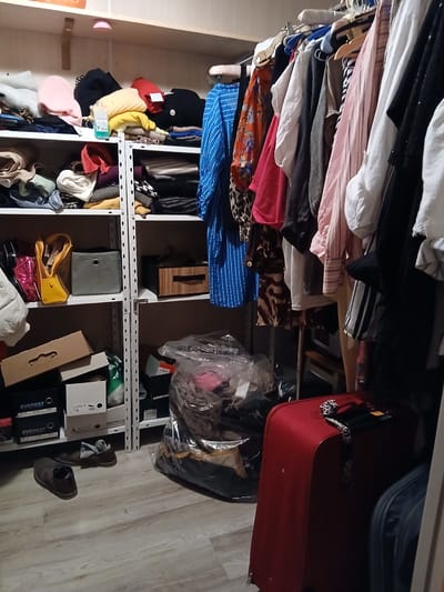 The image depicts an indoor scene, likely a closet or storage room, filled with various items. On shelves, there are neatly folded clothes, including sweaters and tops in different colors. Among the clothing, there are also bags, boxes, and a gray fabric storage cube. On the floor, there are several pairs of shoes, including a pair of brown leather shoes. To the right of the shelves, a large red suitcase is visible, and to its right, a portion of another dark-colored suitcase is present. Hanging clothes are on a rack to the right of the shelves, showcasing a variety of shirts, dresses, and jackets. The environment appears to be a residential or personal storage space. The location is identified as Vita-Poshtova, Ukraine.