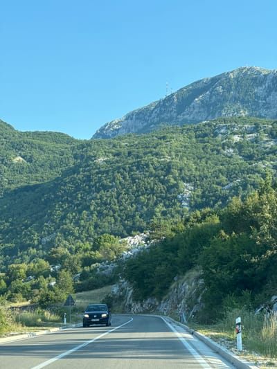 In the city of Kopito, Montenegro, a black sedan is driving on a paved road that curves to the right. The road has white lane markings. The car is in the right lane. On the left side of the road, there is a triangular warning sign on a post. The road is surrounded by lush green vegetation and rocky terrain. In the background, a large, majestic mountain rises, covered in dense trees and topped with rocky outcrops and several telecommunication towers. The sky above is a clear, vibrant blue.