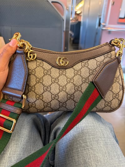 A person is seated inside a passenger train car in Linden, United States, holding a Gucci Ophidia GG mini bag. The bag, resting on the individual's lap, features a beige GG Supreme canvas pattern with brown leather trim and a prominent gold-toned interlocking GG logo on the front. A short brown leather handle with gold-toned chain hardware is held by the person's right hand, which displays a darker skin tone. A wider shoulder strap, characterized by green and red Web stripes with brown leather accents and gold-toned buckles, drapes across the person's light blue denim-clad legs. The train's interior background is visible, showing a light gray overhead luggage rack, elongated fluorescent ceiling lights, and reflections on interior surfaces, indicating a clean and modern public transport environment.