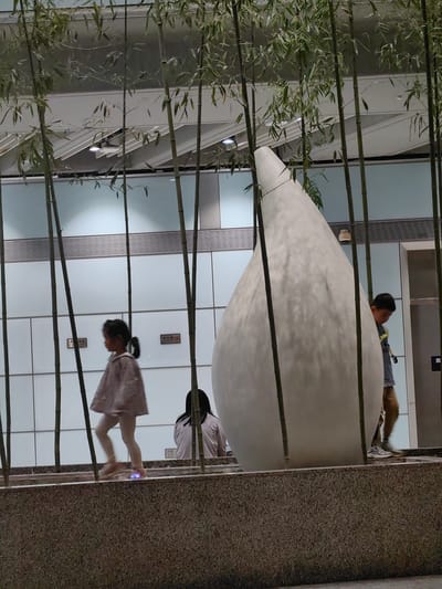 The image shows an indoor scene with bamboo stalks and a large, white, teardrop-shaped sculpture. A young girl wearing a light jacket and leggings walks past the sculpture from left to right. Behind the sculpture, a person is sitting, and to the right of the sculpture, a boy walks from right to left. The background features a wall with horizontal lines and panels. A concrete base supports the bamboo and sculpture. The location is likely in a public space or building within the Shunyi District, China.