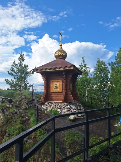 A small, wooden, Orthodox chapel with a golden dome stands on a rocky outcrop. The chapel features an icon in a gold frame on one side. A metal railing surrounds the area, leading towards the chapel. The background consists of lush green trees and rolling hills under a bright blue sky with scattered white clouds. The location is the "Skala Belaya" (White Rock) natural park in Sim, Russia.