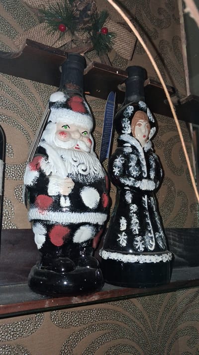The image shows two decorative bottles on a shelf. The bottle on the left is shaped like Santa Claus, with a white beard, red cheeks, and a black suit with red and white polka dots. The bottle on the right is shaped like a woman in a black coat with white snowflake patterns and a fur-lined hood. Both bottles have black tops. Behind the bottles, a wooden shelf is visible, adorned with pine branches and a pine cone. The background consists of a patterned brown wallpaper. The image is taken at a slight angle, with the left bottle being closer to the viewer. No people are present in the image. The scene appears to be indoors, possibly in a home or shop, decorated for the Christmas season. The location of Marchevo, Bulgaria is not discernible from the image itself.