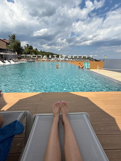 This image depicts an outdoor leisure complex with an infinity pool in Hlibivka, Ukraine. The scene is observed from the perspective of a person reclining on a light grey sun lounger, with their bare legs and feet, showing light pink toenails, prominently in the foreground.

The midground features a large, clear blue infinity pool. Several individuals are present in the pool: at least two are partially submerged on the left side, and one person is lounging near the right edge, adjacent to two upright blue and white paddleboards. Along the left edge of the pool, multiple white sun loungers and shaded cabanas are visible, extending towards a multi-story building with a light brown facade. A light-colored platform with additional white sun loungers and umbrellas lines the far side of the pool. A volleyball net is partially visible at the far right edge of the pool area.

The background reveals a vast, calm body of water extending to the horizon, indicative of a river or reservoir. A Ukrainian flag, blue and yellow, flies from a flagpole on the far right, overlooking the water. Green trees and foliage line a distant bank or hill on the left side. The sky is partially cloudy, with large white and grey clouds against a blue backdrop, casting distinct shadows on the wooden decking in the foreground.