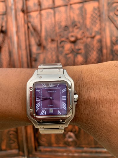 A close-up shot captures a person's arm wearing a Cartier Santos wristwatch. The arm, with medium to dark brown skin tone and visible fine hair, is positioned against a blurred, ornate background. The background appears to be a textured, reddish-brown surface, possibly a decorative metallic gate or wooden panel, featuring intricate, swirling patterns.
The wristwatch is the focal point, showcasing a distinctive square-shaped silver-toned case with prominent screws on the bezel. Its striking purple dial features white Roman numeral hour markers and a date window at the 4 o'clock position, displaying "18". Visible text on the dial includes "CARTIER" positioned centrally above the watch hands and "QUARTZ" towards the bottom right, indicating its movement type. The watch is attached to a matching silver-toned bracelet, also secured with visible screws. Minor scratches are visible on the watch case and bracelet, suggesting it has been regularly worn. The overall impression is a casual display of a luxury timepiece. The scene is set in Ijeododo, Nigeria.
