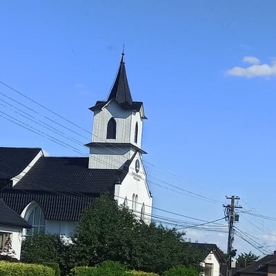 The image shows a white church with a black, pointed steeple against a clear blue sky. The church has vertical siding on the tower and a black tiled roof. There are several sets of overhead power lines in front of the church. In the foreground, there are green bushes and hedges, and in the background, there are other buildings and trees. The church appears to be located in a residential area. The text on the church reads "Biserica Baptistă" and "Milișăuți". This indicates the church is a Baptist church in the town of Milișăuți, Romania.
