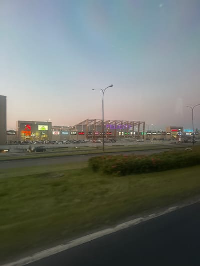 The image captures a dusk scene of the Felicity shopping mall in Lublin, Poland, as viewed from a moving vehicle. The facade of the mall is illuminated with prominent signage, including "CINEMA CITY," "LEROY MERLIN," "H&M," "PRIMARK," "CCC," and "RESERVED," indicating a diverse retail offering. The mall features a modern architectural design with a wooden-slatted roof structure. Numerous vehicles are visible in the parking lot. A road runs in front of the mall, with green grass and some flowering plants lining the median. The sky is a soft gradient of light blue and pink, characteristic of twilight. Streetlights are also visible along the road.