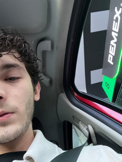 The image shows a person sitting inside a car at a PEMEX gas station. The person is looking upwards and has curly dark hair and a short beard. A seatbelt is visible. The car's sunroof is open, and the PEMEX sign is visible above. The sign indicates "8" on a green background. The location is San Francisco Totimehuacán, Mexico.