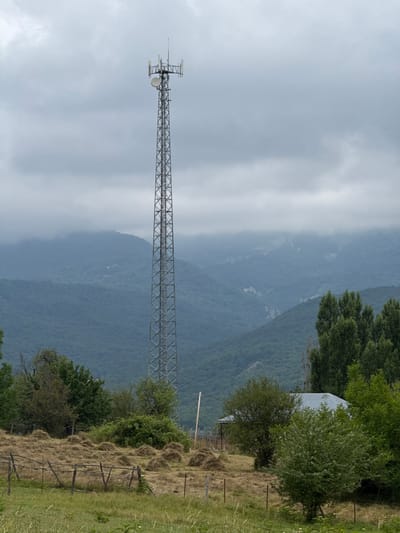 The image shows a telecommunications tower in a rural setting, with mountains in the background under a cloudy sky. In the foreground, there is a field with haystacks and a rustic wooden fence. A small building with a metal roof is partially visible among trees. The overall scene suggests a remote, possibly agricultural area. The location provided in the prompt, Sis, Azerbaijan, places this scene within that geographical context. No people or specific actions are depicted.