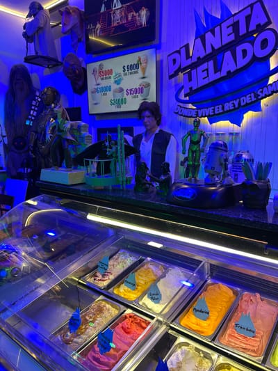 The image captures the interior of "Planeta Helado," a Star Wars-themed ice cream shop in Villa General Belgrano, Argentina, illuminated by an intense blue light that gives the space a futuristic ambiance.
In the middle ground, a person, likely an employee, is visible behind a dark counter, attending to something below eye level. The shop is heavily decorated with an array of Star Wars characters and props. Various figures and statues are displayed, including multiple R2-D2 units, C-3PO, Darth Vader, Chewbacca, Yoda, stormtroopers, and other alien creatures, creating an immersive experience for visitors.
To the right, a brightly lit glass display case showcases numerous colorful ice cream flavors in metal tubs. Visible flavor labels include "Frambuesa" (Raspberry), "Mandarina" (Mandarin), "Limon Polar" (Polar Lemon), "Mascarpone con Frutos Rojos" (Mascarpone with Red Berries), "Maracuya" (Passion Fruit), "Banano Split" (Banana Split), and "Frutilla a la Crema" (Strawberry Cream).
Above the counter, a digital screen displays the pricing: "$5000 UNA BOCHA" (one scoop), "$9000 LAS 2 BOCHAS" (two scoops), "$10000 ½ KILO", and "$24000 1 KILO". A prominent blue sign on the back wall reads "PLANETA HELADO" (Ice Cream Planet) and "DONDE VIVE EL REY DEL SAM" (Where the King of Sam lives). The artificial lighting suggests the time of day is evening or night.