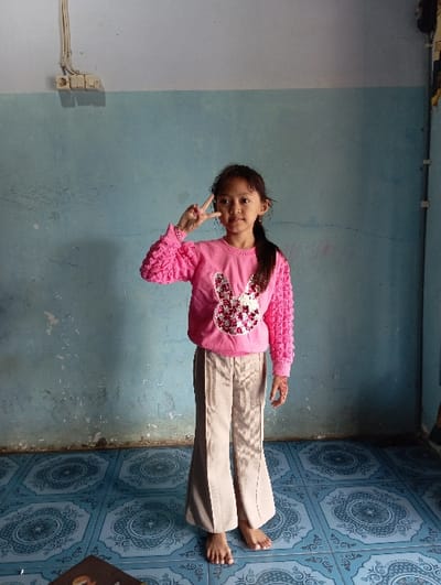 The image depicts an indoor scene in Bulukandang, Indonesia, featuring a single young female standing barefoot and facing the viewer directly. She has dark hair styled in a ponytail over her left shoulder and exhibits a neutral facial expression while making a peace sign (V-sign) with her raised right hand. On her left hand, an intricate reddish-brown design, consistent with a henna tattoo, is visible. She wears a long-sleeved pink sweater with textured, possibly ruffled or bobble-knit sleeves, and a large, sequined bunny rabbit motif on the chest. A light pink beaded bracelet is present on her right wrist. She is also wearing light brown or beige wide-leg trousers.
The background consists of a plain light blue wall, exhibiting some visible scuff marks and discoloration, particularly in the lower sections. The upper portion of the wall, above a horizontal line, is painted a lighter shade, likely off-white or very pale blue. In the upper left quadrant of the light blue wall, an exposed electrical conduit system is visible, featuring three horizontally aligned white electrical outlets or switches connected by a vertical conduit pipe. The floor is covered with blue and white square tiles, each featuring a repeating ornate circular or medallion-like pattern. A small portion of an unidentified dark object is visible in the extreme bottom left corner of the frame.