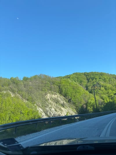 From the vantage point of what appears to be a moving vehicle, the image captures a roadside scene. A highway, marked by double white lines, runs alongside a verdant, tree-covered hillside. Utility poles with wires traverse the slope, interspersed among the trees. The sky is a clear, unblemished blue, with the faintest crescent moon visible in the lower left quadrant. A metal guardrail separates the road from the hillside. The overall impression is one of a natural landscape juxtaposed with transportation infrastructure, likely observed while traveling through a mountainous area. The location in the city of Echinos, Greece, is not explicitly evident from the scene alone.
