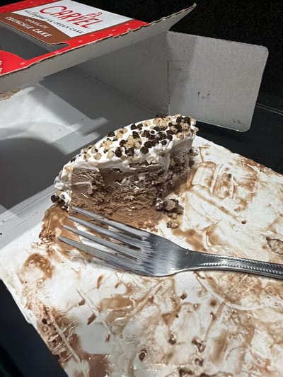 A partially eaten Carvel Chocolate Crunchie Cake is on display inside its open cardboard box. The cake, which features layers of chocolate ice cream and crunchies, has been served on a white tray that is now smeared with chocolate frosting and ice cream drippings. A silver fork rests on the tray, its tines embedded in the remaining cake. The box itself is branded with the Carvel logo and product name. The scene is dimly lit, suggesting it may be nighttime or indoors in low light. No individuals are visible in the image. The location is identified as Wildwood, United States.