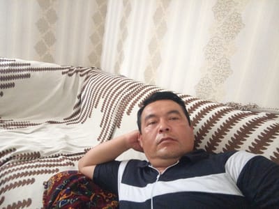 The image shows a man lying on a couch with his head propped up on his hand. The couch is covered with a white and brown patterned blanket. Behind the couch, there is a light-colored wall with a subtle textured pattern. The man is wearing a black and white striped polo shirt. The setting appears to be an indoor living space. The image quality is typical of a selfie taken with a mobile phone. There are no specific identifiable landmarks or details that would place this image in Hakkulabad, Uzbekistan.