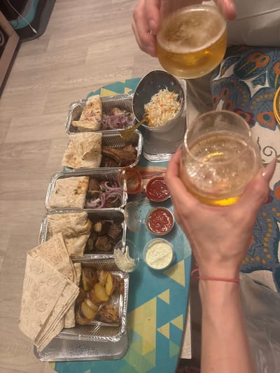 Two people are toasting with glasses of beer. They are seated at a table with a colorful, patterned tablecloth. In front of them are four foil trays containing food: one tray has a salad of shredded cabbage and carrots, and the other three trays contain grilled meats, flatbread, and roasted potatoes. Several small containers of sauces or dips are also visible. The scene appears to be indoors, possibly a home setting, with a wooden floor. The lighting suggests it is daytime. The overall mood seems relaxed and social, with people enjoying a meal and drinks. There is no visible text in the image.