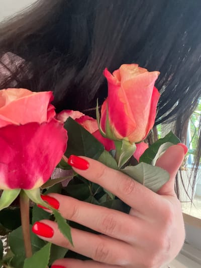In this close-up, vertically oriented image, a person's hand with bright red fingernails is holding a bouquet of two prominent roses, one primarily red and the other a gradient of pink and orange. The hand is positioned in the lower half of the frame, obscuring a portion of the roses. The person's long, dark hair is visible in the upper portion of the image, framing the bouquet and hand. Behind the roses, a softly blurred background suggests an indoor setting with natural light filtering in, possibly from a window. The roses themselves are in focus, showcasing their layered petals and green stems and leaves. The image appears to have been taken in Bayreuth, Germany, though no specific landmarks or contextual clues within the image itself pinpoint a precise location within the city.