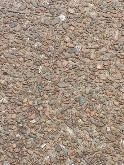 The scene depicts a ground surface in Konongo, Ghana. The surface is composed primarily of small, irregularly shaped pebbles and gravel, ranging from approximately 0.5 cm to 4 cm in maximum dimension, set within a lighter, granular matrix of tan or light brown material. The pebbles exhibit a variety of natural earth tones, including shades of brown, reddish-brown, grey, and off-white. Scattered among the pebbles and matrix are numerous small, fragmented pieces of white and off-white debris, some appearing to be shell fragments due to their curved or irregular shapes, while others are elongated or angular. No individuals, animals, significant manufactured objects, specific actions, or interactions are present in the frame. The surface is rough and uneven, illuminated by what appears to be natural daylight.