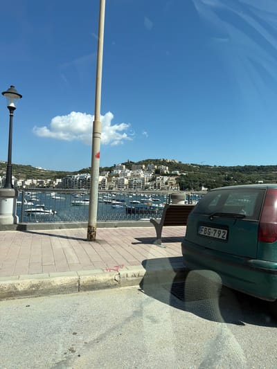 The image shows a view of the bay and coastline of Saint Paul's Bay, Malta, on a clear, sunny day. In the foreground, a green Fiat Punto car is parked on the right side of the frame, with its license plate visible as FBG 792. To the left of the car is a paved walkway with a concrete curb and a metal railing along the edge of the bay. A street lamp stands on the walkway, and further left, a concrete pillar with the number 75 is visible.
The bay itself is filled with numerous boats, mostly small to medium-sized vessels, at anchor. The water is a deep blue, reflecting the clear sky above. Beyond the bay, the town of Saint Paul's Bay sprawls up a hillside, characterized by its densely packed white and light-colored buildings. The hill is covered in lush green vegetation. The sky is a vibrant blue with a few scattered white clouds. The overall scene depicts a typical Mediterranean coastal town.