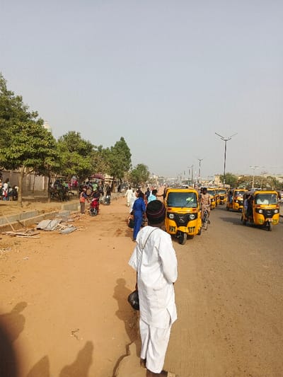 The image depicts a daytime street scene in Gobirawa, Nigeria. In the immediate foreground, a person is observed from behind, wearing a white traditional tunic and trousers, with a dark knitted cap and a dark bag slung over their left shoulder. They stand on an unpaved, reddish-brown dirt shoulder.

To the left, the dirt shoulder features scattered construction debris, including concrete slabs and wooden planks. Further back, several green-leafed trees line a low wall or barrier. Beyond these trees, a cluster of individuals is visible, some standing, some seated, near what appears to be market stalls or informal structures. A red motorcycle is parked near the shoulder, with a person in an orange safety vest nearby.

To the right, a multi-lane paved road extends into the distance. A procession of yellow three-wheeled vehicles, commonly known as keke-napep or autorickshaws, occupies the right side of the road. The closest vehicle displays the license plate "MAY 0777". Several individuals are present on or near the paved road, including a person in blue overalls and another riding a bicycle.

The background shows the road receding, flanked by modern streetlights with dark poles and multi-fixture heads. The sky is a pale, hazy blue-grey. The overall environment suggests a busy urban or semi-urban area with ongoing activity and a mix of pedestrian and vehicular traffic.