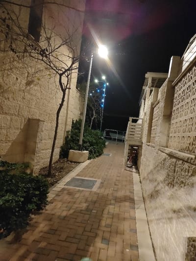 A narrow brick pathway stretches between stone buildings under the night sky. A streetlamp illuminates the path. A bare tree and green bushes flank the left side of the path, while a building with patterned walls borders the right. A bicycle is nestled beneath a staircase on the right side of the path, leading towards a distant street with parked cars and strings of blue lights. The scene captures a quiet urban night.