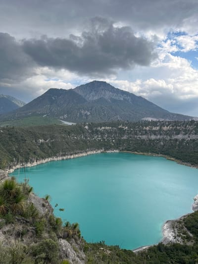 A high-angle, wide shot captures the Crater Lake of San Luis Atexcac, Mexico, a striking body of turquoise water nestled within a volcanic crater. The lake's vibrant color contrasts with the rugged, grey and green slopes of the crater rim, which is covered in dense vegetation. In the background, a large, imposing mountain range dominates the horizon, its peaks shrouded in a dramatic sky of heavy, dark clouds interspersed with patches of bright blue. The foreground features rocky, arid terrain with sparse, spiky green plants. The scene is devoid of any people or visible activity, presenting a serene and natural landscape.
