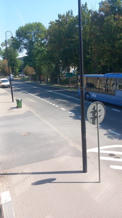 The image depicts a street scene in Góra Kalwaria, Poland, on a sunny day. The foreground is dominated by a paved sidewalk with a light gray tiled surface. A large, dark gray shadow of a lamppost stretches across the sidewalk from the right. To the left, a green trash bin with wheels is situated near the edge of the sidewalk. Beyond the sidewalk, a multi-lane asphalt road is visible, marked with white dashed lines in the center. A white van is parked on the left side of the road, and a blue bus with the "nka" logo is traveling in the right lane. In the background, numerous tall green trees line the street, and a blue sky with faint white contrails is overhead. A street signpost with a circular sign featuring a cross-like symbol is visible on the right side of the road.