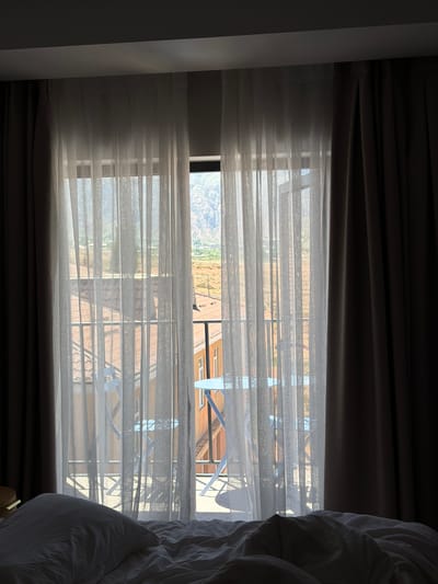 The image shows a bedroom with a bed in the foreground and a view through sheer curtains of a balcony and a mountainous landscape outside. The bed is covered with white sheets and a pillow. The sheer curtains are white and hang from a rod, obscuring some of the view but allowing light to filter through. On the balcony, there are two blue chairs and a table. Beyond the balcony, there are buildings with red-tiled roofs and then a rugged, mountainous area in the distance. The light suggests it is daytime, with sunlight streaming through the window and casting shadows. The scene appears to be located in Kaji-Say, Kyrgyzstan, a town known for its proximity to Issyk-Kul Lake and surrounding mountains, consistent with the visual elements.