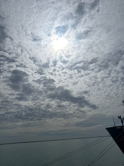 The image captures a coastal scene in Uribia, Colombia, under an overcast sky during daytime. The upper two-thirds of the frame are filled with a dense formation of altocumulus clouds, through which the sun is visible as a bright, diffused light source in the upper center, creating a distinct glare. The cloud cover exhibits varying densities and textures, with darker, more pronounced cloud masses contrasting with lighter areas around the sun.
The lower third of the image features a calm body of water, likely a bay or sea, reflecting the grey tones of the sky. Two small, red-topped navigational buoys are present on the water's surface: one in the lower-left quadrant and another in the lower-right, closer to the distant horizon. A low-lying, linear landmass or coastline stretches across the horizon, separating the water from the sky.
In the lower-right corner, the dark-colored bow of a large, stationary vessel is prominently visible, extending vertically into the frame. Multiple thick mooring lines originate from the vessel and descend diagonally towards the bottom of the image. A portion of the ship's superstructure, including a mast or antenna with attached fixtures, is also visible above the bow. No individuals are present in the depicted scene.