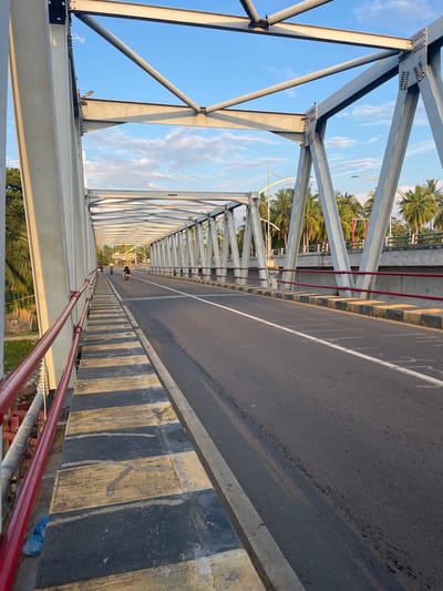 This is a photograph of a bridge in Neuboknaleung, Indonesia. The bridge has a steel truss structure painted white and a red railing. The road on the bridge is asphalt with white lane markings. There are some vehicles, including two motorcycles, on the bridge. The surrounding environment includes palm trees, vegetation, and a blue sky with clouds. The lighting suggests it is either sunrise or sunset.