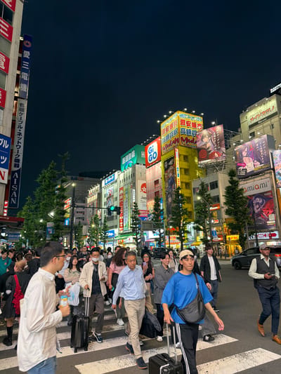 The image depicts a busy street scene at night in Chiyoda, Japan, likely Akihabara, known for its electronic stores and anime culture. A multi-story building prominently features numerous illuminated advertisements and signage in Japanese. Pedestrians are crossing a street on a zebra crossing, some carrying luggage. One individual in the foreground is wearing a blue t-shirt and black track pants with white stripes, pulling a black suitcase. Another person to the right is dressed in a light blue button-down shirt and tan pants, also with a suitcase. The street is lined with trees, and traffic lights are visible. The overall atmosphere is that of a vibrant urban environment at night.