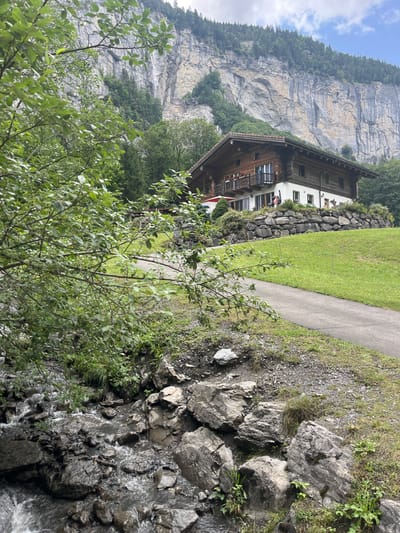 The image depicts a picturesque natural scene in Lauterbrunnen, Switzerland, characterized by lush greenery and dramatic geological formations. In the foreground, a rocky stream flows over dark, wet stones, with branches of green foliage extending from the left side of the frame, partially obscuring the view. A paved pathway winds its way through a manicured green lawn, leading towards a traditional Swiss chalet. The chalet, constructed primarily of wood with white stucco accents, sits on a terraced hillside. A stone wall, constructed from large, irregular rocks, supports the lawn in front of the chalet. Above the chalet, a towering, sheer cliff face rises majestically, its striated surface a testament to immense geological time. A thin waterfall cascades down the cliff face in the distance, barely visible through the trees. The sky above is partly cloudy, with patches of blue peeking through. No people are visible in the scene, and the overall atmosphere is one of serene, unspoiled natural beauty.