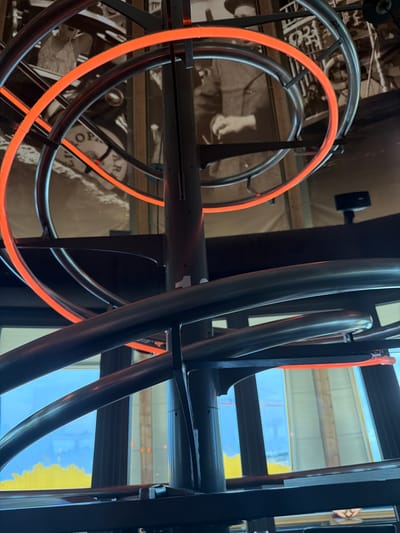 The image shows an abstract close-up of a spiral staircase with neon orange lighting. The staircase is made of dark metal pipes. In the background, there is a sepia-toned mural depicting historical figures. The scene appears to be indoors, with windows visible in the lower part of the image, reflecting a bright, possibly yellow, outdoor landscape and a blue sky. The overall impression is one of modern design incorporated into a historical setting. While the image is striking, it does not contain enough information to determine a specific location within the city of Rust, Germany.