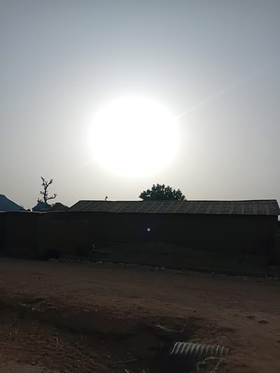 The image depicts a rural or semi-rural scene in Nata-Ala, Nigeria, under direct sunlight. The prominent feature is an overexposed white sun centrally located in a hazy, light gray sky, creating strong backlighting. The foreground consists of reddish-brown dirt ground with visible textures, some scattered organic debris resembling twigs, and small white fragments. In the lower right corner, a partially buried corrugated metal culvert extends into the frame. The midground is dominated by a long, horizontal, dark-silhouetted structure, likely a building, constructed from what appears to be earthen material, topped with a dark, corrugated metal roof. A small, indistinct dark figure is visible on the peak of this roof, slightly to the left of its center. In the background, behind the main structure, several silhouetted trees are present: a gnarled, bare-branched tree to the left, and a dense, leafy green tree further right. Another partial roofline is visible to the far left. No discernible human figures are clearly identifiable. The overall environment suggests a dry, outdoor setting.