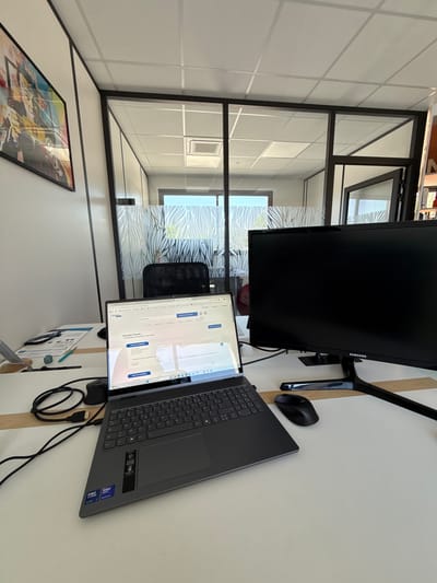 The image depicts an indoor office setting in Les Arcs, France. A desk occupies the foreground, hosting a laptop, a Samsung monitor, and a mouse. A chair is visible behind the laptop screen. A framed artwork hangs on the wall, beneath a windowed partition wall, which is translucent. The environment beyond the partition wall shows office spaces, and the ceiling is composed of square panels.