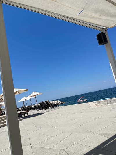 The image shows a beach scene in Crveni Vrh, Croatia. The foreground features a white paved walkway with a light gray concrete floor. To the left, a white pillar and an overhead white canopy with a rip in it frame the view. A black speaker is mounted on a white post under the canopy.

In the middle ground, several white beach umbrellas are set up over lounge chairs arranged on a pebble beach. A few people are visible lounging on the chairs, one with their legs extended. The sea stretches out in the background, a deep blue under a clear sky. A small, red and white watercraft, resembling a pedal boat, is floating on the water. Further out, there are some buoys and distant landmass visible on the horizon. The overall impression is a sunny, serene beach resort environment.