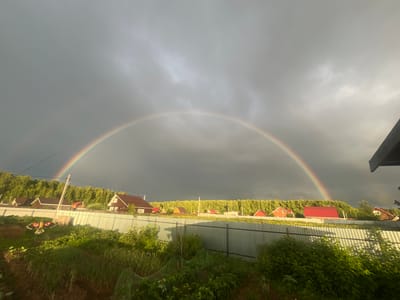 This is a wide shot of a rural landscape in Klepiki, Russia, featuring a vibrant double rainbow arching across a dramatic, cloudy sky. The scene is set in what appears to be a suburban or dacha community, with a row of houses visible behind a corrugated metal fence. Lush green vegetation, including what looks like a garden and trees, fills the foreground and middle ground, suggesting it's late spring or summer.
The sky is a mix of dark, stormy clouds on the left and brighter, sunlit clouds on the right, indicating a recent or passing shower. The sunlight breaking through creates the spectacular rainbow phenomenon. The houses have red roofs and wooden exteriors, typical of Russian countryside homes. In the foreground, there are garden beds with various plants and what appears to be a scarecrow or decorative kite shaped like a bird of prey.
The time of day is likely late afternoon or early evening, given the quality of light and the contrast between the dark clouds and the illuminated areas. The overall mood is one of natural beauty and perhaps a sense of calm after a storm. There is no visible text in the image.